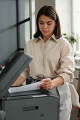 Fototapete - Young female office manager standing by xerox machine and putting paper on copier glass while taking copies of financial document