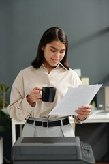Fototapete - Happy young businesswoman with financial papers and cup of coffee standing by xerox machine in front of camera and reading documents
