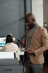 Fototapete - Young African American businessman using smartphone and tablet while standing in front of xerox machine and making copies of documents