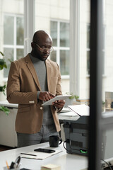 Wall Mural - Young confident businessman in formalwear pointing at tablet screen while networking in front of xerox machine with copies of paper documents