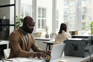 Fototapete - Young serious white collar worker typing on laptop keyboard while sitting by workplace with xerox machine and working over new project