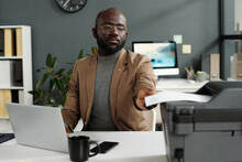 Young African American Male Employee With Laptop Taking Printed Paper Document From Tray Of Xerox Machine While Sitting By Workplace