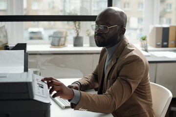 Fototapete - Young office worker pressing start button on xerox panel to copy document while sitting by his workplace and typing on keyboard