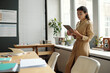 © pressmaster - Young serious woman in quiet luxury attire watching online video on tablet screen while standing by window in front of desk with copybooks