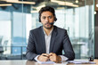 © Liubomir - Close-up photo of a serious young, Indian male businessman sitting in the office, at a desk in front of the camera and wearing a headset.