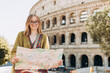 A happy blond woman tourist with paper map is standing near the Coliseum, old ruins at the center of Rome, Italy. Concept of traveling famous landmarks. 30s Girl is walking on a sunny day