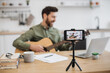 © sofiko14 - Skillful musician sitting at home office using smartphone and tripod for filming video tutorial about guitar playing. Handsome male blogger sharing with followers in social media artistic experience.