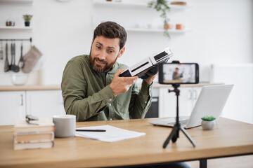 Wall Mural - Caucasian surprised male creating online content on wireless smartphone for personal channel. Shocked man telling about modern technologies and brand new robot vacuum cleaner in living room interior.