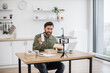 © sofiko14 - Positive handsome man recording video blog while unboxing new modern smartphone indoors. Interesting influencer leaving review about product and sitting at table with laptop in room.