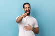 Well you're a joker. Portrait of positive handsome man with beard wearing white T-shirt pointing index finger at camera and laughing positively. Indoor studio shot isolated on blue background.