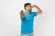 Portrait of disgusted handsome unshaven man wearing blue T- shirt standing and pinching nose, smelling awful odor, showing stop gesture. Indoor studio shot isolated on gray background.