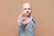 © khosrork - Portrait of bald bearded man does stop sign with palm, demonstrates prohibition and restriction, refuses something, says no, wearing gray jacket. Indoor studio shot isolated on brown background.