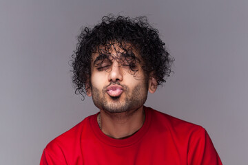 Portrait of romantic flirting handsome bearded man with curly hair, standing with closed eyes, keeps lips pout, sending kissing. wearing red jumper. Indoor studio shot isolated on gray background.