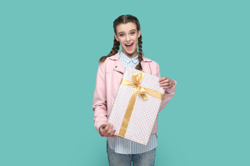 Wall Mural - Portrait of excited positive teenager girl wearing pink jacket standing wrapped present box in hands, looking at camera, waiting holiday to give gift. Indoor studio shot isolated on green background.