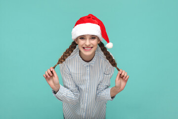 Wall Mural - Portrait of funny optimistic teenager girl with braids wearing striped shirt and Santa Claus hat, looking at camera and pulling her pigtails aside. Indoor studio shot isolated on green background.