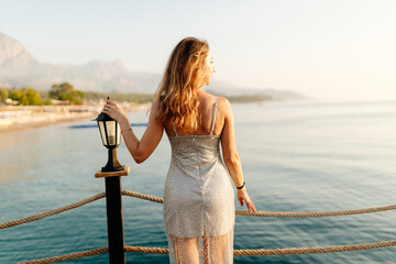 Wall Mural - A slender girl with long hair in a gray shiny dress walks along the pier by the sea at sunset. A beautiful woman in an evening dress poses against the backdrop of the ocean at dawn. Summer background