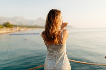 Wall Mural - A slender girl with long hair in a gray shiny dress walks along the pier by the sea at sunset. A beautiful woman in an evening dress poses against the backdrop of the ocean at dawn. Summer background