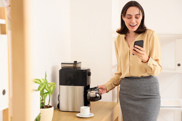 Canvas Print - Beautiful young businesswoman using mobile phone while making coffee in office