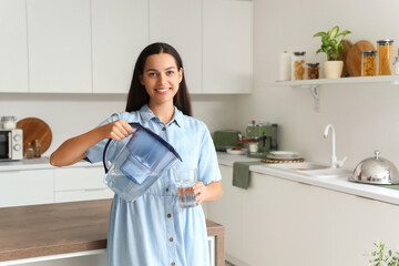 Poster - Beautiful young woman pouring pure water from filter jug into glass in kitchen