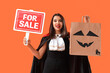 © Pixel-Shot - Young woman dressed for Halloween with gift bag and FOR SALE sign on orange background