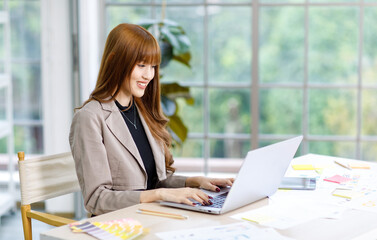 Asian professional successful young female businesswoman creative graphic designer employee in casual fashionable suit blazer outfit sitting working using typing laptop computer at office workstation