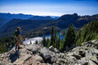 Adventurous athletic male hiker standing on top of a hill taking a photo of an alpine lake and a rugged mountain range on a beautiful sunny fall day in the Pacific Northwest.
