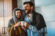© NDABCREATIVITY - Happy young couple with hot drinks cuddling under warm blanket at home