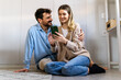 © NDABCREATIVITY - Young happy couple using phone to share social media news at home, doing shopping online.