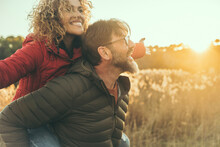 Happy Couple Have Fun Together In Outdoor Leisure Activity In Nature Field During Sunset Time And Golden Hours Light. Man Carry Woman On His Back. People Enjoying Life And Laughing. Winter Autumn Day