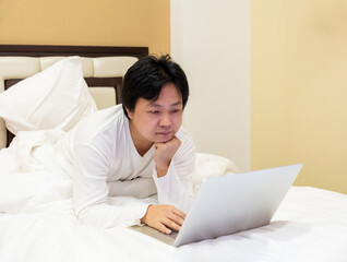 Work from home, a freelancer man leaning against the bed and using his laptop to work online on a bed.