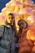 © Glittering Humanity - Two women, bundled in beautiful puffy coats, stand in the winter landscape, their portraits glowing in the neon lights as they brave the outdoor chill with their stylish jackets whimsy from a mushroo