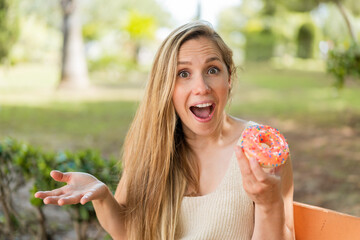 Wall Mural - Young blonde woman holding a donut at outdoors with shocked facial expression