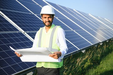 Wall Mural - Portrait of Young indian man technician wearing white hard hat standing near solar panels against blue sky.Industrial worker solar system installation, renewable green energy generation concept.