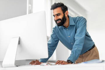 Smiling indian male entrepreneur looking at computer monitor, businessman standing at workplace in office