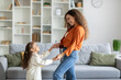Happy young mother and daughter dancing and having fun at home, child girl and her mommy spending weekend together, side view, copy space
