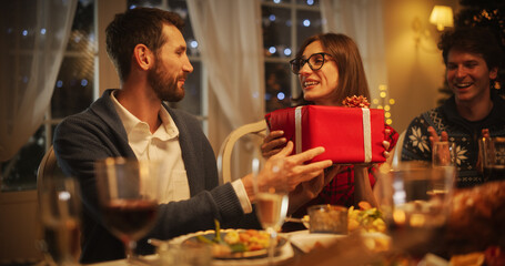 Wall Mural - Handsome Boyfriend Presenting a Festive Gift to His Beautiful Surprised Girlfriend. Family and Friends Gather at Home in the Evening for a Traditional Christmas Dinner with a Turkey Roast Feast