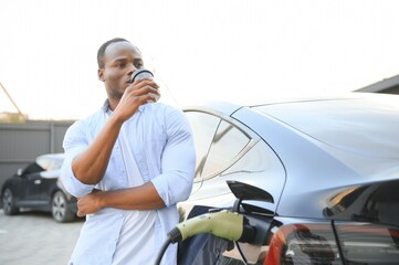 African man standing near electric car with charging cable in plug. Eco friendly vehicle charging on station. LIfestyles concept