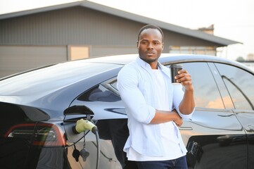 African man standing near electric car with charging cable in plug. Eco friendly vehicle charging on station. LIfestyles concept