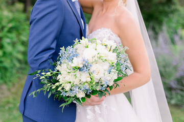 Wall Mural - A bride and groom are standing together. The bride wears a white wedding dress and long veil, and she holds a bouquet of white flowers with greenery and touches of blue flowers.