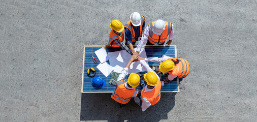 Top view of engineers team, male and female put hands on top over blueprint, solar photovoltaic equipment, road and wind turbine business important infrastructure on solar panel at construction site.