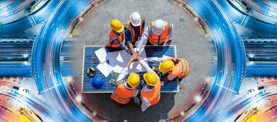 Top view of engineers team, male and female put hands on top over blueprint, solar photovoltaic equipment, road and wind turbine business important infrastructure on solar panel at construction site.