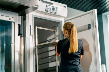 Shot from the back of a girl a pastry chef puts candy to harden in the refrigerator and sets cooling temperature