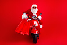 Portrait Of Excited Cool Santa Claus With White Beard In Sunglass Driving Scooter With Shopping Bags Isolated On Red Color Background