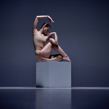 Like A Statue. Elegant Young Woman, Professional Ballerina In Beige Bodysuit Posing On Cube Against Blue Studio Background. Concept Of Classical Dance, Art And Grace, Beauty, Choreography, Inspiration