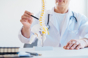 Closeup no face medical male doctor pointing on human spine model with a pen sitting in his office. Spinal healthcare and back pain treatment concept. Selective focus, copy space