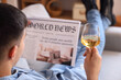 Young man with glass of wine reading newspaper at home, closeup