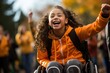 A schoolgirl in a wheelchair, wearing an orange jacket, is happy and laughing on the street while supporting someone with a raised hand.
