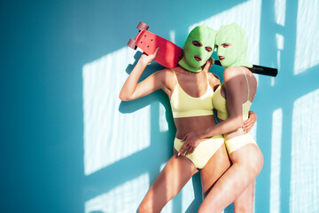 Two beautiful sexy women in green underwear. Models wearing bandit balaclava mask. Hot seductive female in nice lingerie posing near blue wall in studio. Crime and violence. Hold penny gun skateboard