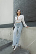 © alones - Stylish beautiful Caucasian woman with sunglasses in fashion urban clothes with a white blazer, vintage high waisted blue jeans and sneakers with a bag stands near a black brick building. Pretty lady