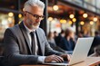 © Georgii - Middle-aged Caucasian man in a suit wearing glasses sits at a table with laptop computer. Positive successful businessman, entrepreneur, small business owner works online. Remote work concept.
